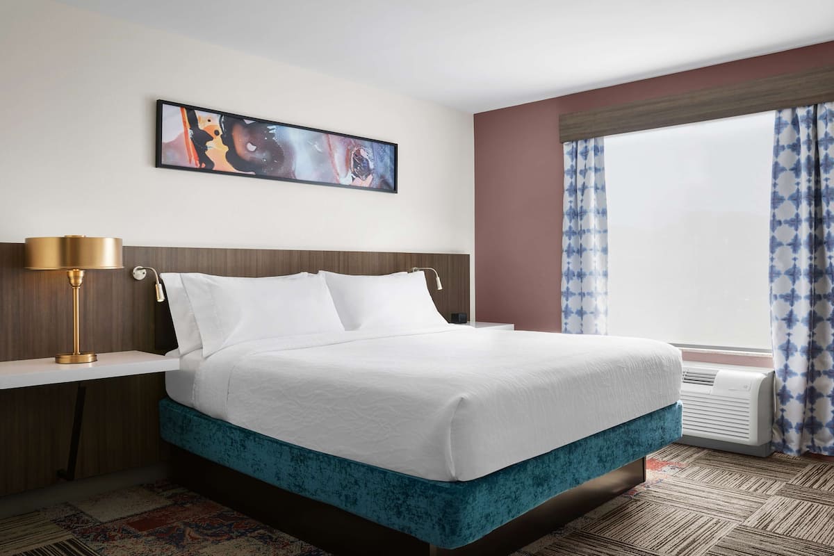 A hotel room with a large bed, a bedside table with a lamp, a wall-mounted painting, and a window with curtains.