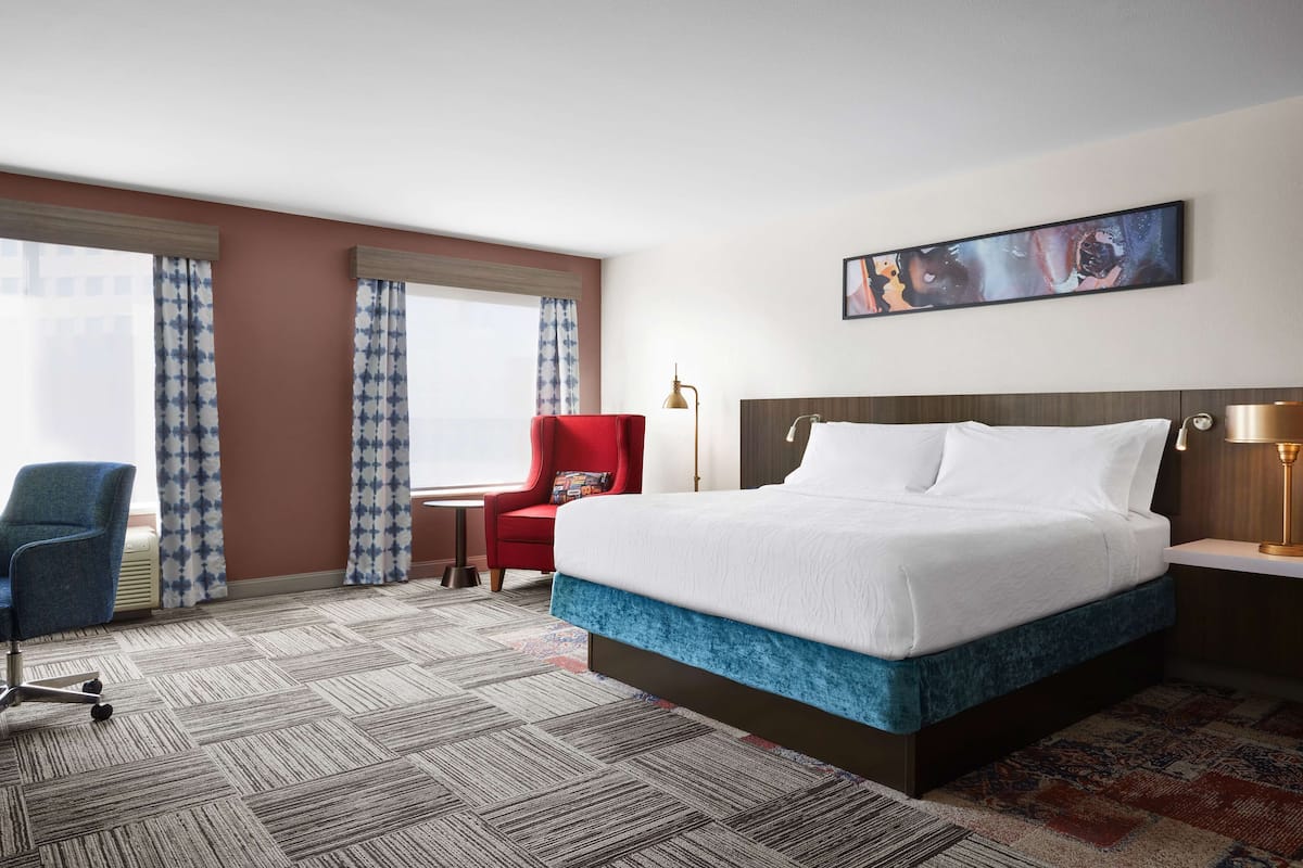 A hotel room with a large bed, a red armchair, a blue office chair, and a small table with a lamp.