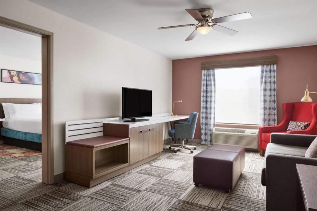 A hotel room with a bed, a desk with a TV, a red armchair, a grey sofa, and a ceiling fan.