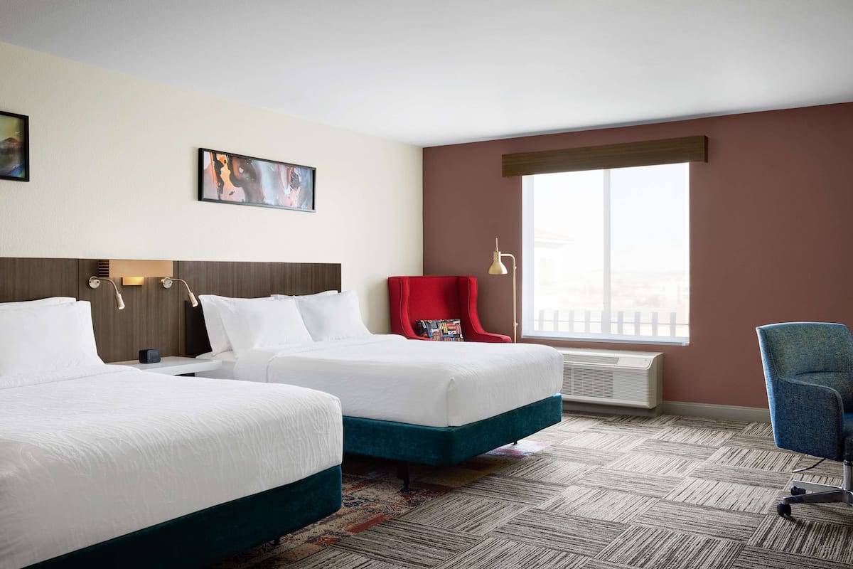 A hotel room with two beds, a red chair, a desk, and a large window.