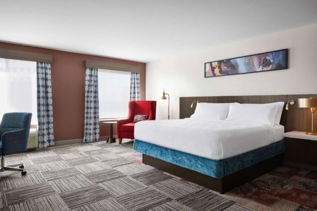 A hotel room with a large bed, a red armchair, a blue office chair, and a patterned carpet.
