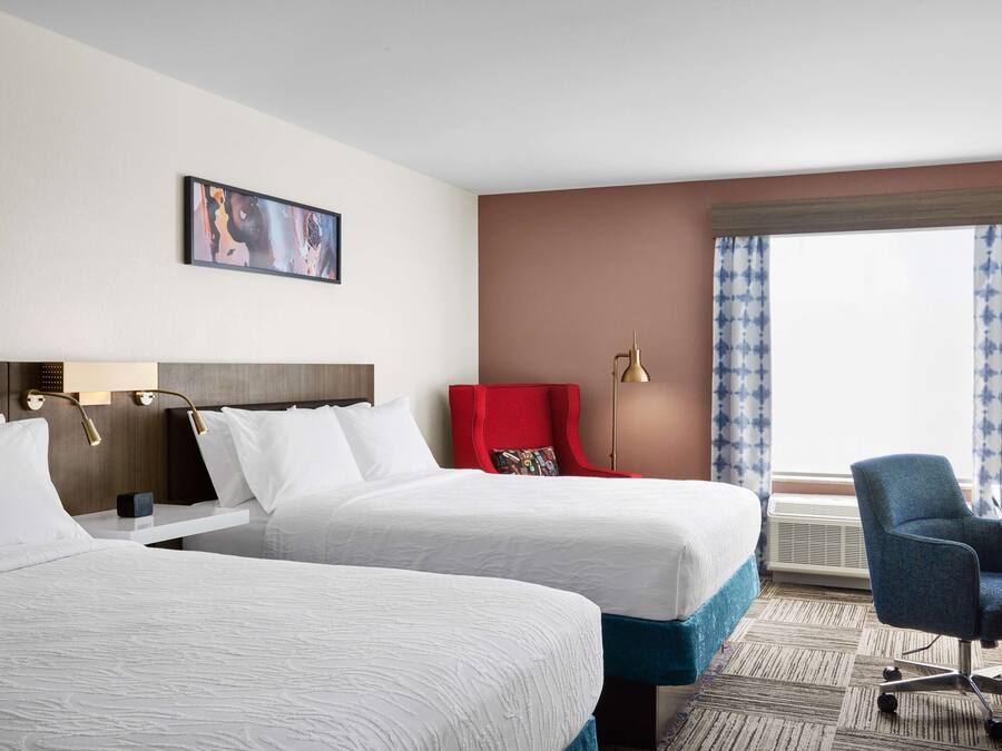 A hotel room with two beds, a red chair, a desk, and a large window with curtains.