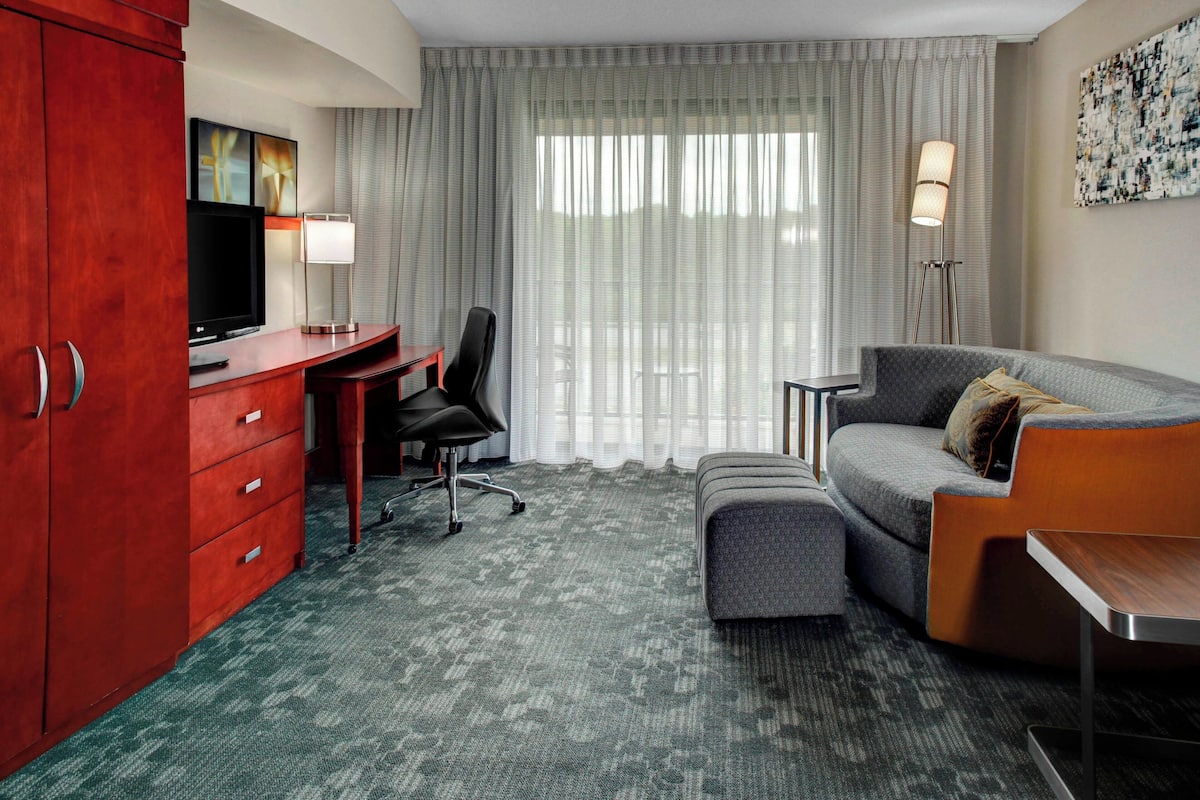 A hotel room with a desk, a grey sofa, a red wardrobe, a television, a floor lamp, and a large window with sheer curtains.