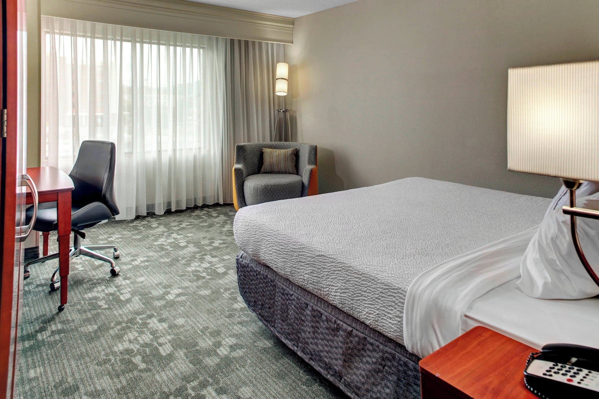 A hotel room with a large bed, a desk with a chair, a lamp, and a view of the outdoors through sheer curtains.