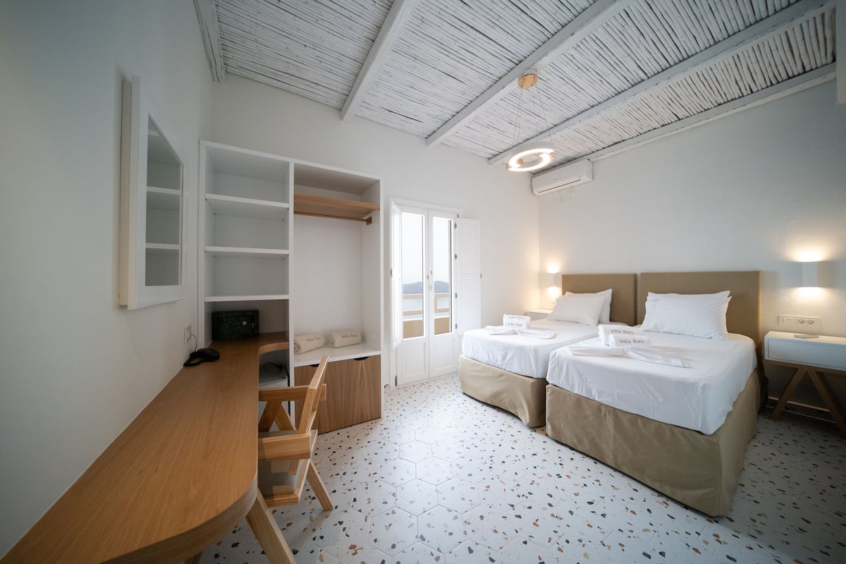 A hotel room with a wooden desk, two beds, a wardrobe, and a large window.