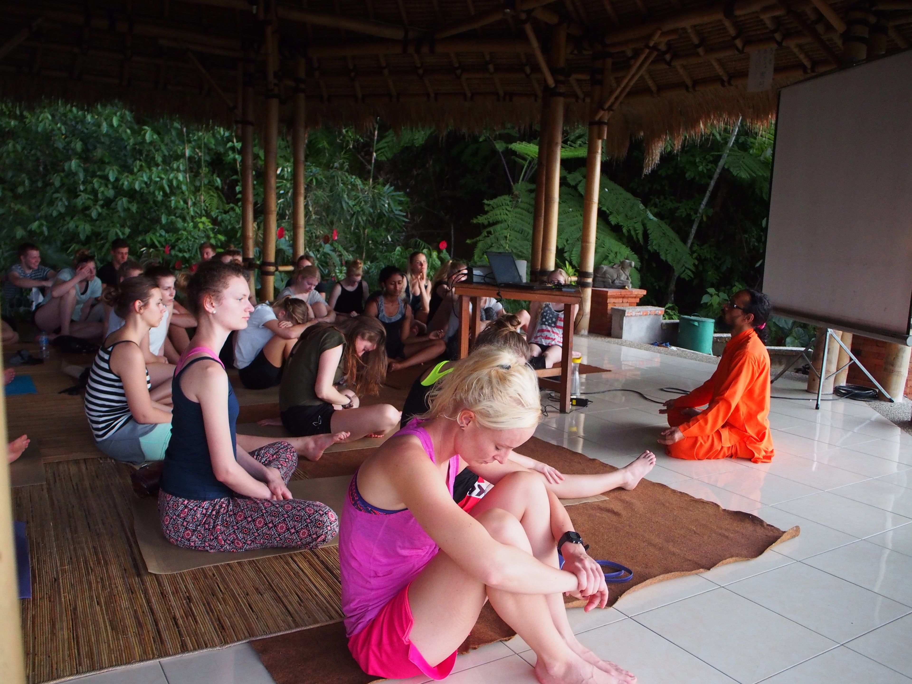 A group of people sitting on mats in a covered outdoor area with a bamboo structure overhead.