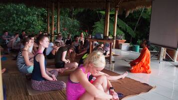 A group of people sitting on mats in a covered outdoor area with a bamboo structure overhead.