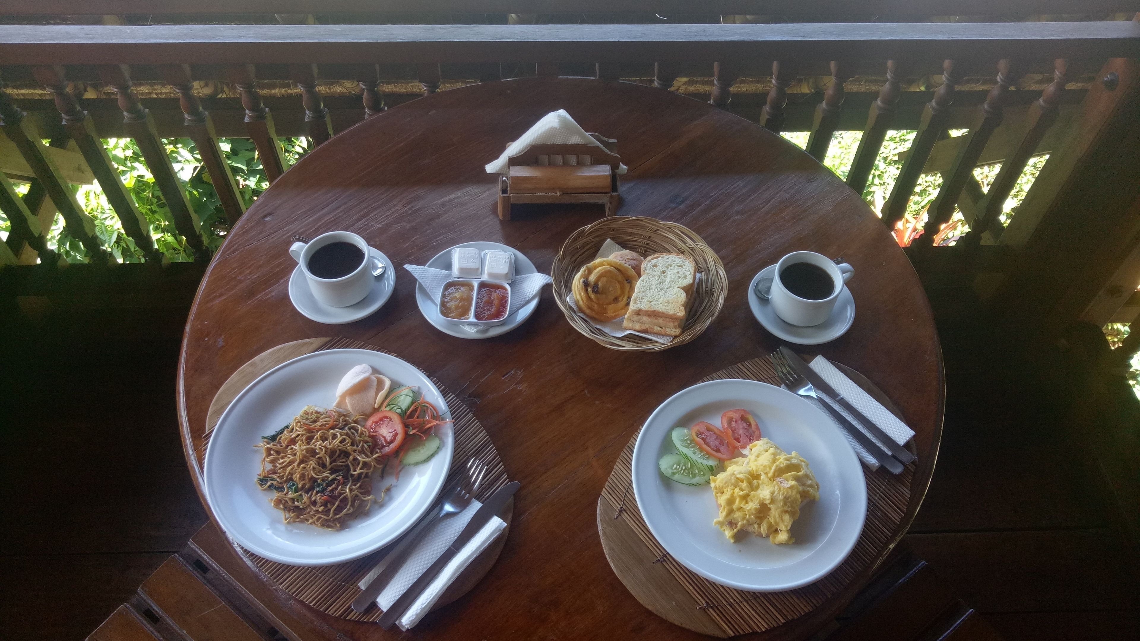 A wooden table with a variety of breakfast items including plates of noodles, scrambled eggs, and sandwiches, along with cups of coffee and condiments.