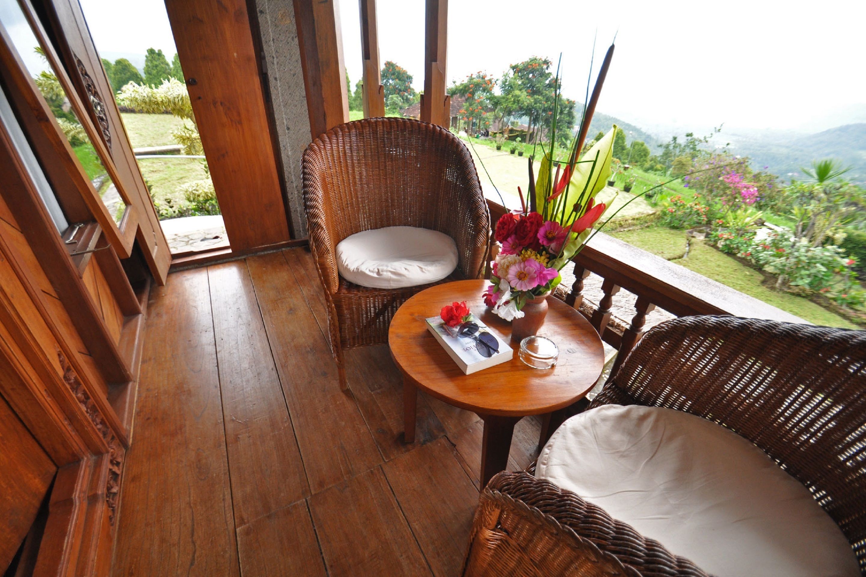 A wooden balcony with wicker chairs, a small table with a vase of flowers, and a view of a lush garden and mountains.