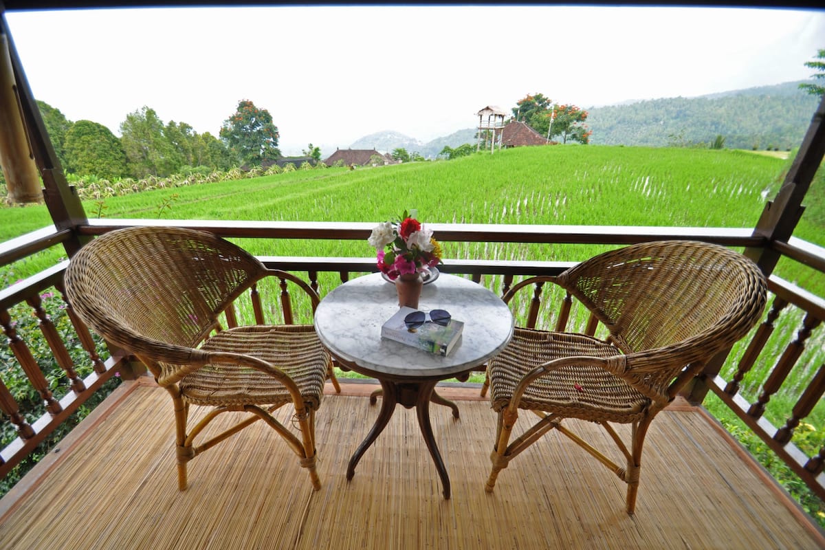 A balcony with wicker chairs and a table, overlooking a green field and distant hills.