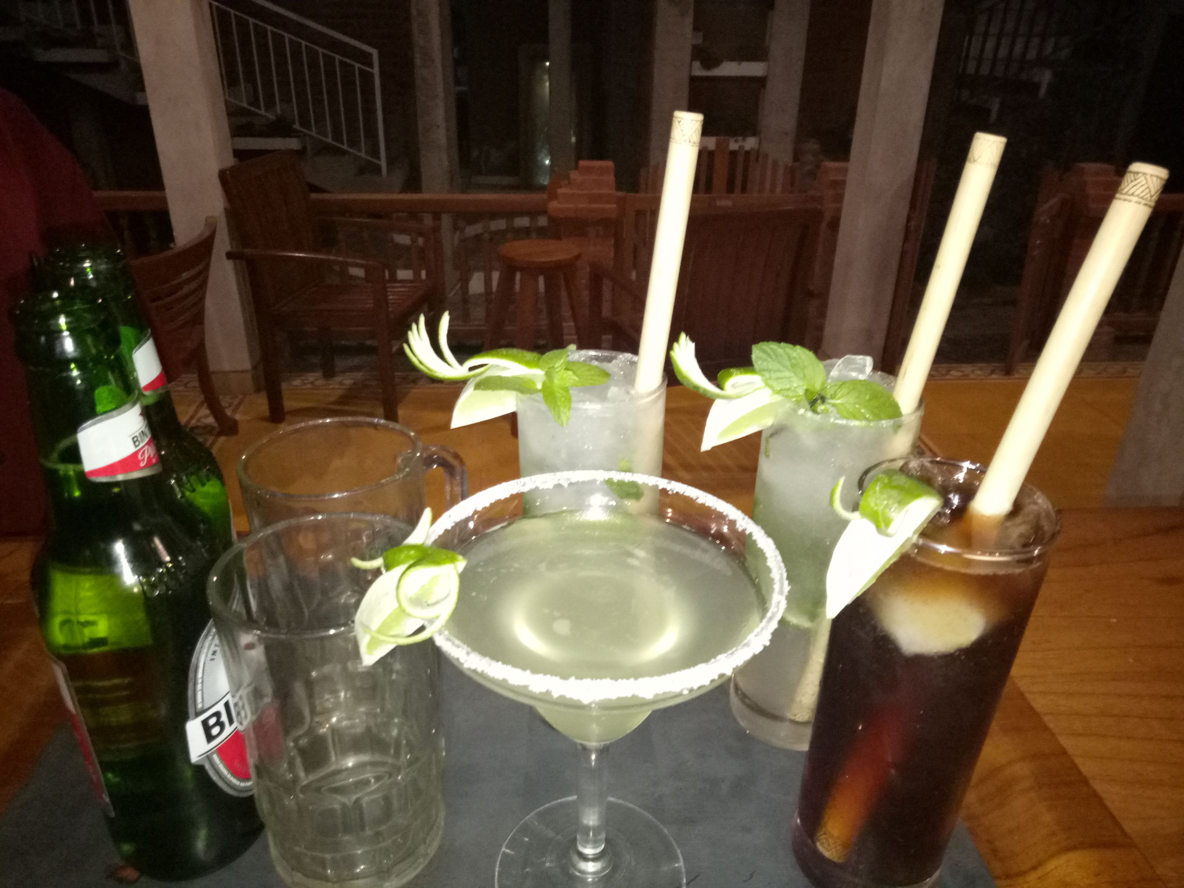 A table with various drinks, including a margarita, a mojito, and a cola, with lime garnishes.