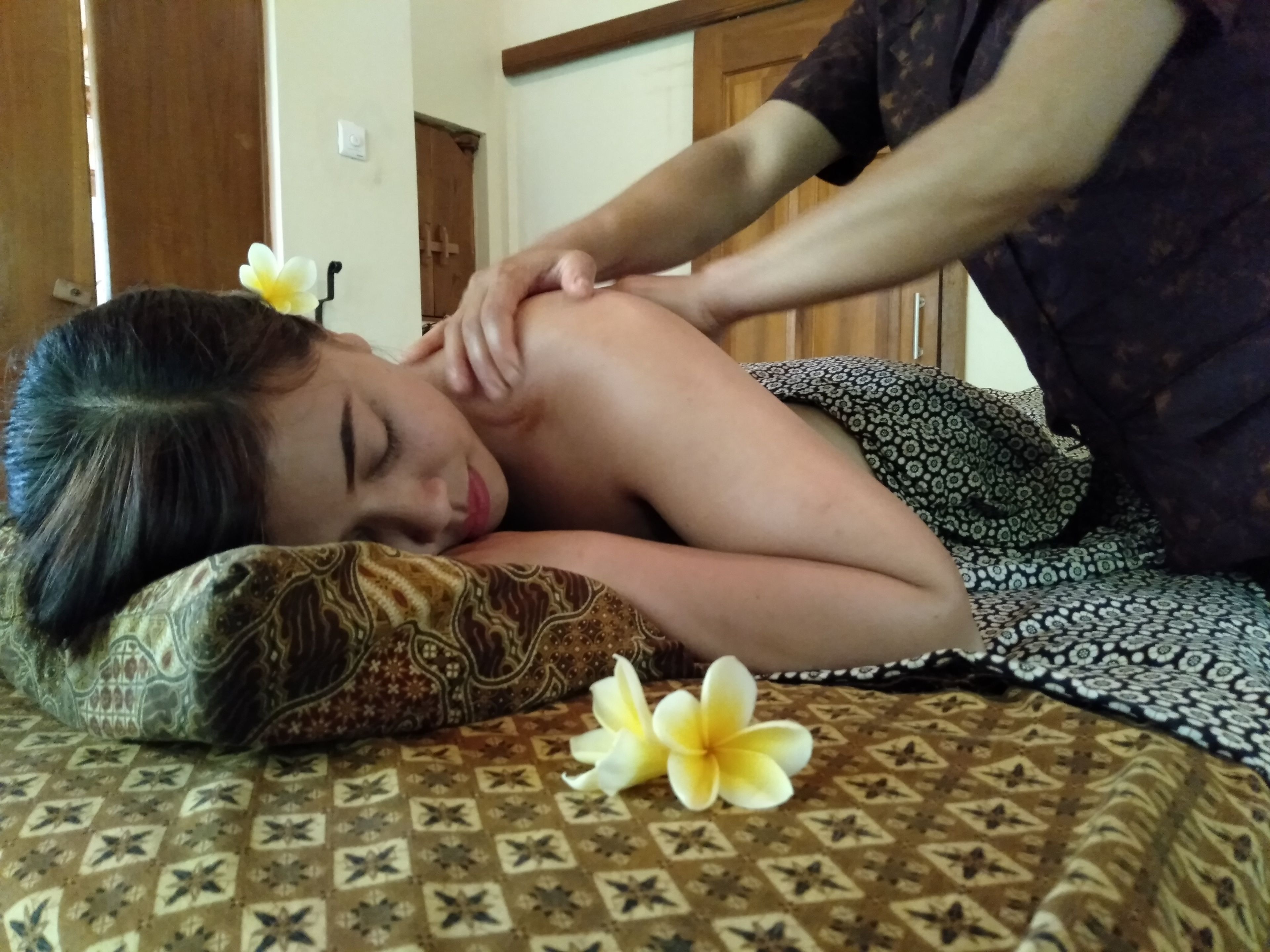 A person receiving a massage on a patterned bed with a pillow and flowers.