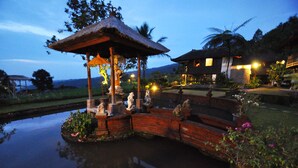 A gazebo with a thatched roof, a pond with statues, and a house with a stone wall in the background. - Puri Lumbung Cottages, Restaurant & Spa (Munduk)
