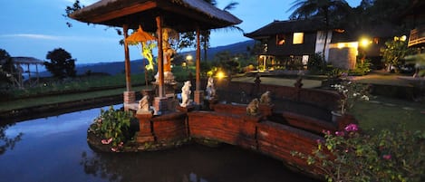 A gazebo with a thatched roof, a pond with statues, and a house with a stone wall in the background.