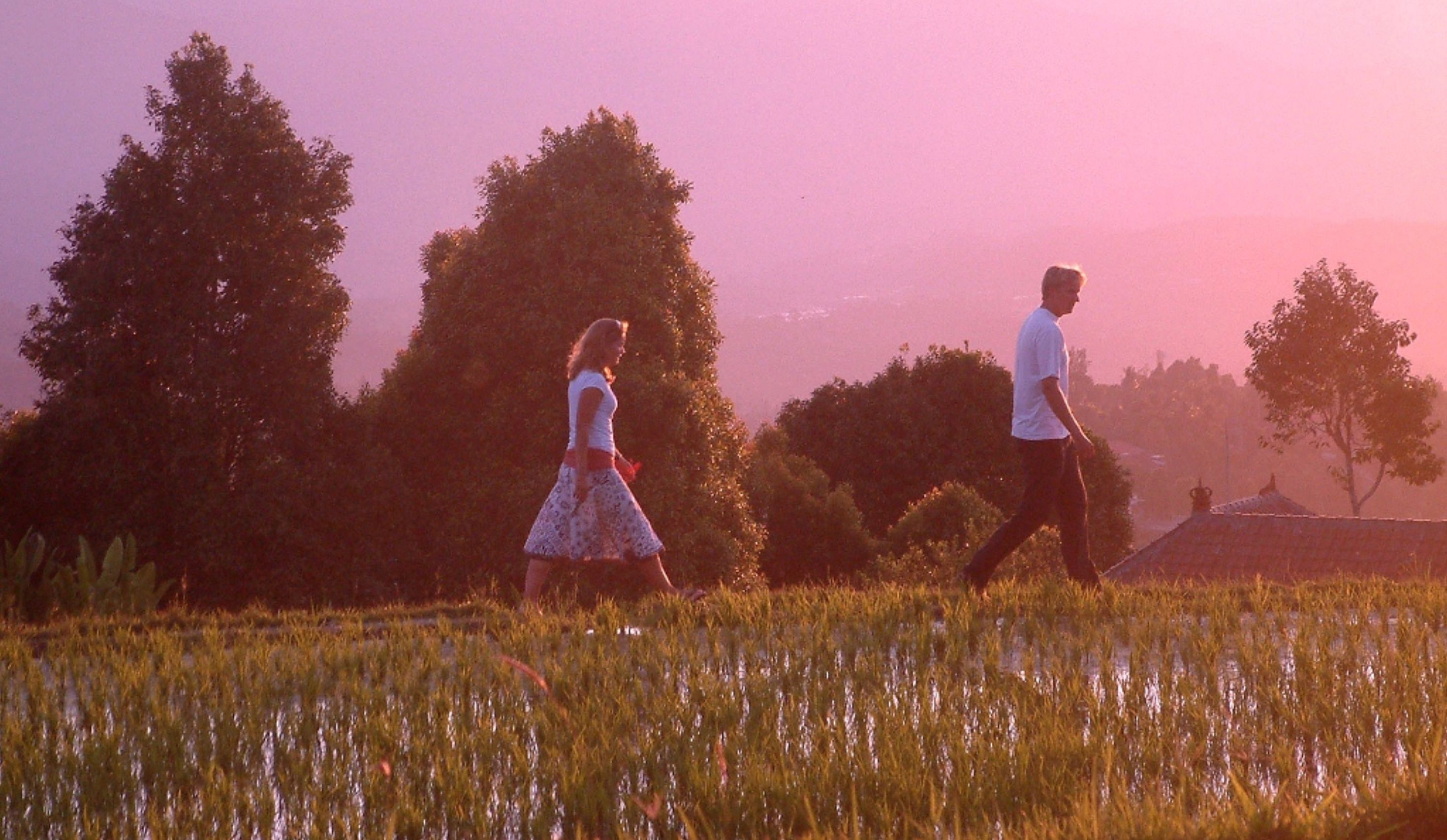 A field with two people walking, trees, and a building in the background.