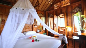 A bed with a canopy, a bedside table with books, a rose, and a potted plant. - Puri Lumbung Cottages, Restaurant & Spa (Munduk)