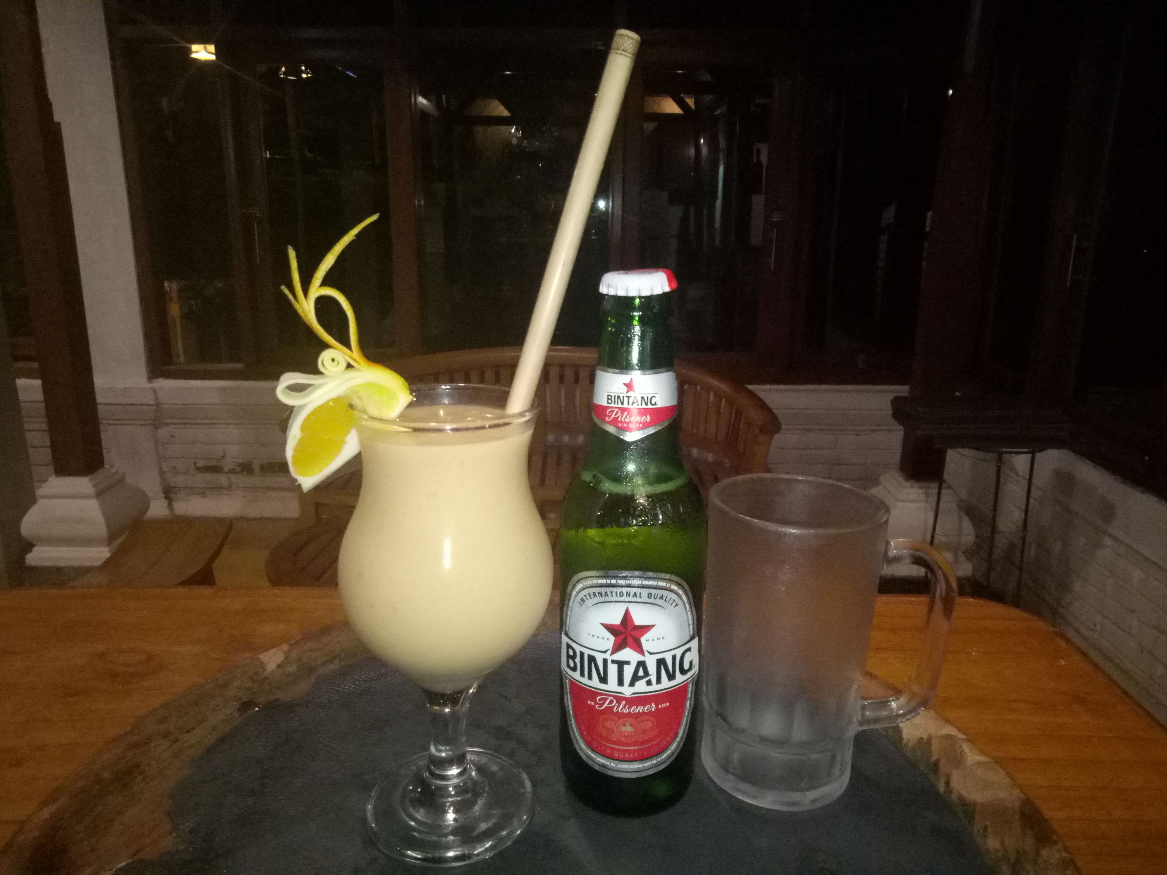 A bottle of Bintang Pilsener beer, a glass of beer with a straw and a lemon slice, and a clear plastic cup on a table.