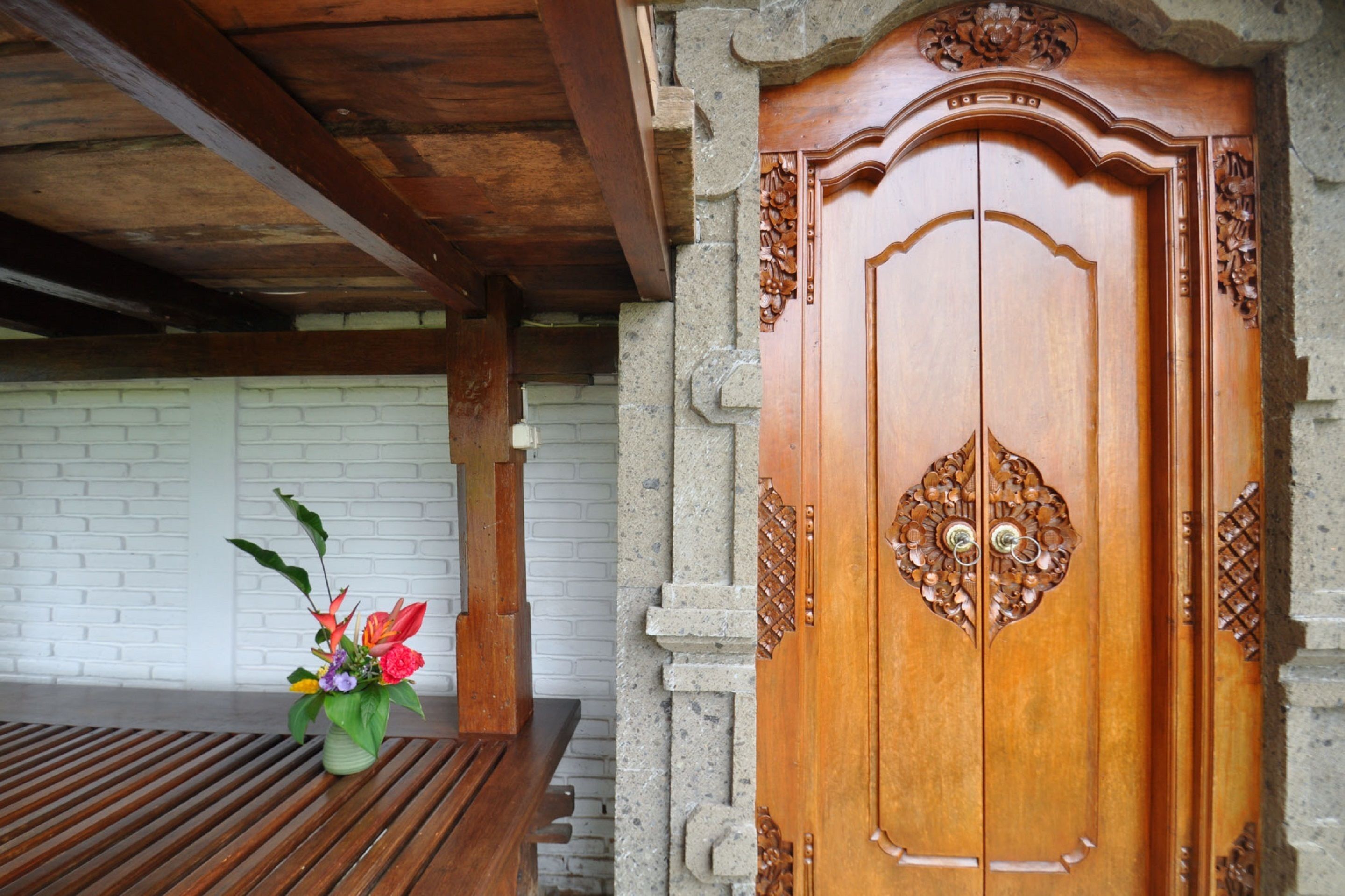A wooden door with intricate carvings, a white brick wall, and a wooden bench with a flower vase.