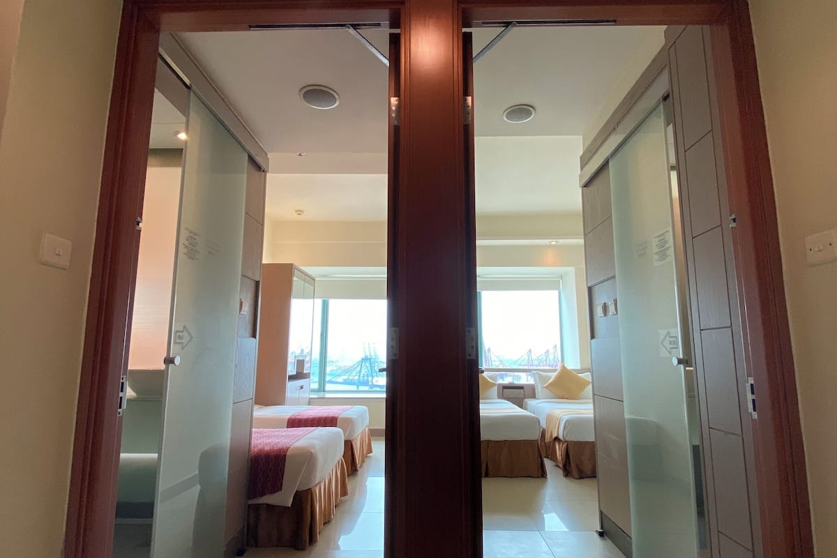 Entrada de hotel com porta de madeira, divisória de vidro e vista para o quarto com camas e janela.