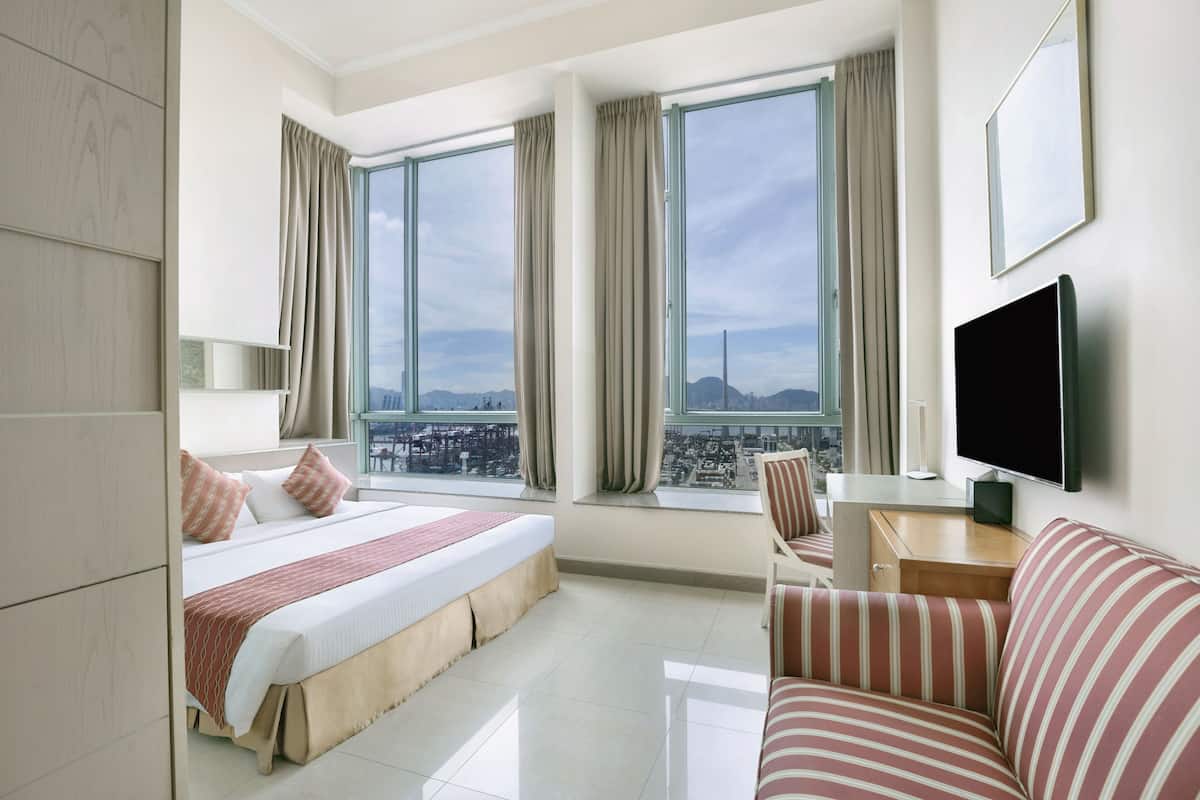 Quarto de hotel com uma cama grande, uma escrivaninha, uma televisão e vista para a cidade através de amplas janelas.