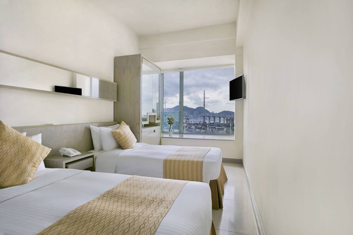 Quarto de hotel com duas camas, uma janela grande com vista para a cidade, televisão montada na parede e telefone ao lado da cama.