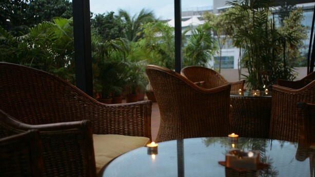 Wicker furniture around a glass table with candles, set in a room with large windows and potted plants. - juSTa OFF MG Road (Bengaluru)
