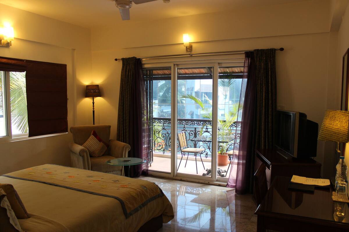 A hotel room with a bed, a TV, a desk, a chair, a table, and a balcony with plants.