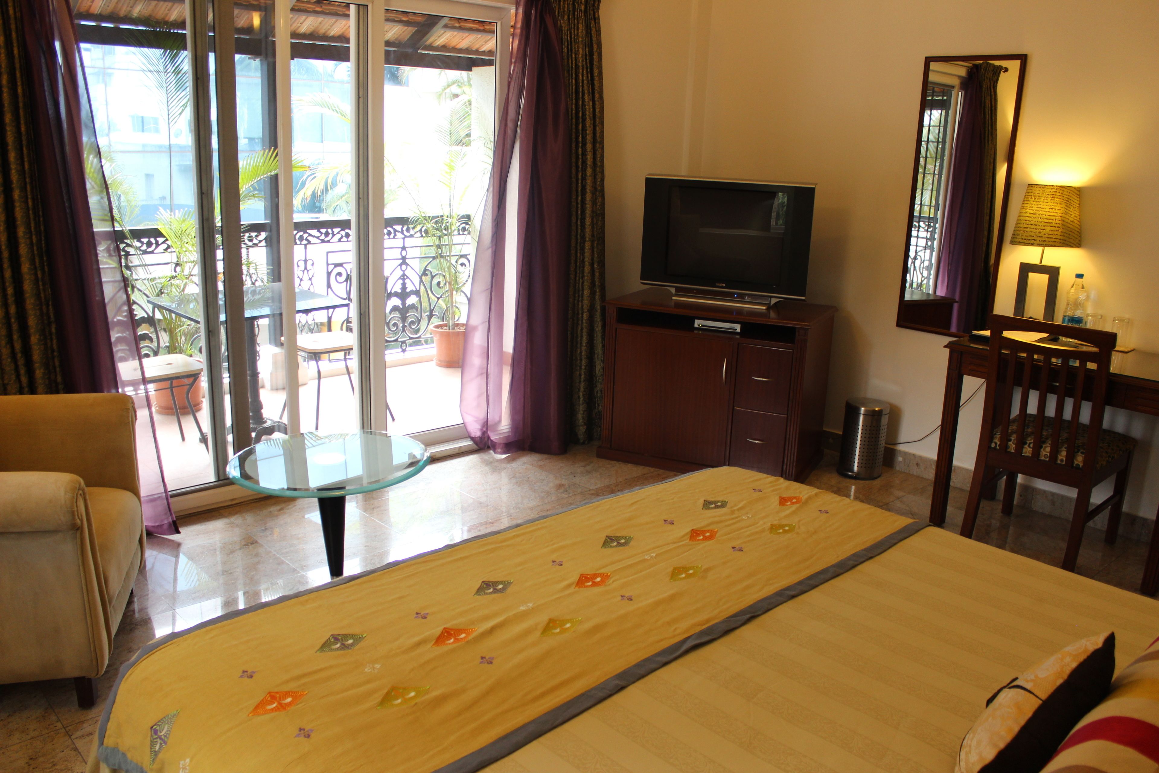A hotel room with a bed, a television, a desk, a chair, and a balcony with plants.