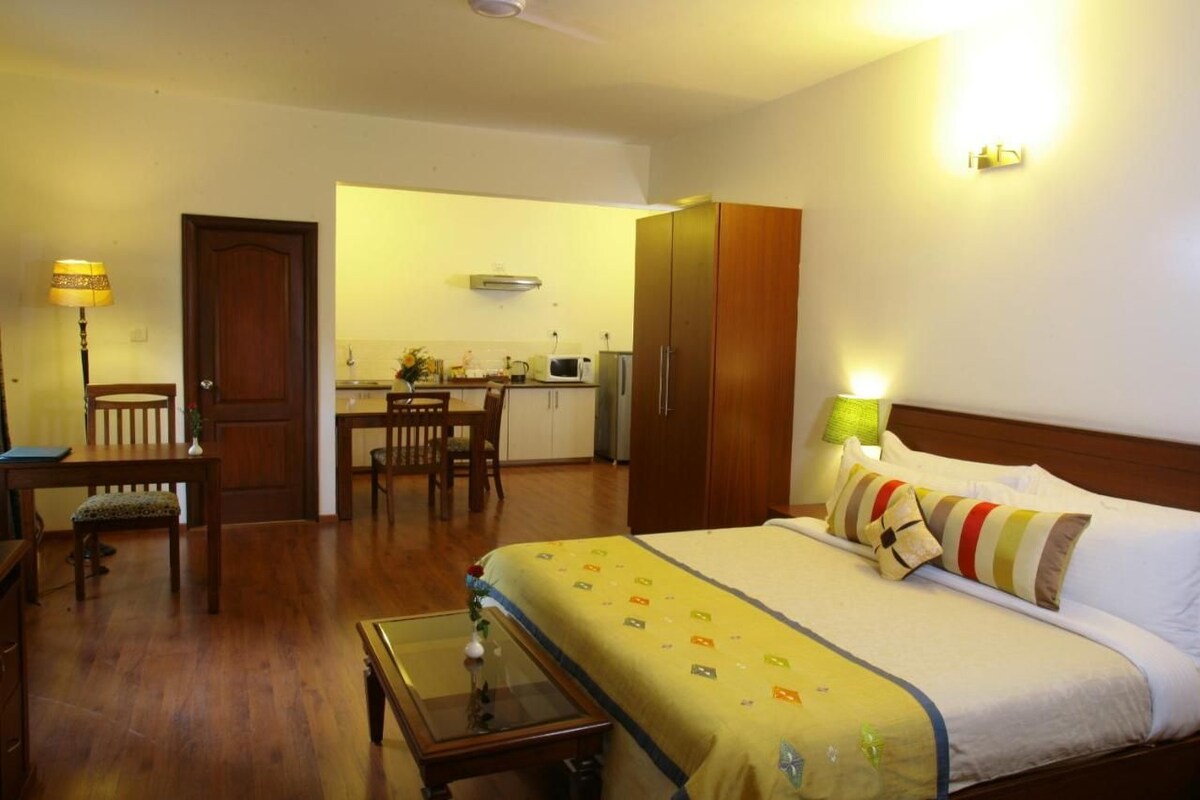 A hotel room with a bed, a desk, a chair, a small table, a lamp, and a kitchenette in the background.