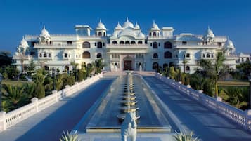 A grand building with a central fountain and a statue, surrounded by a white fence and palm trees.