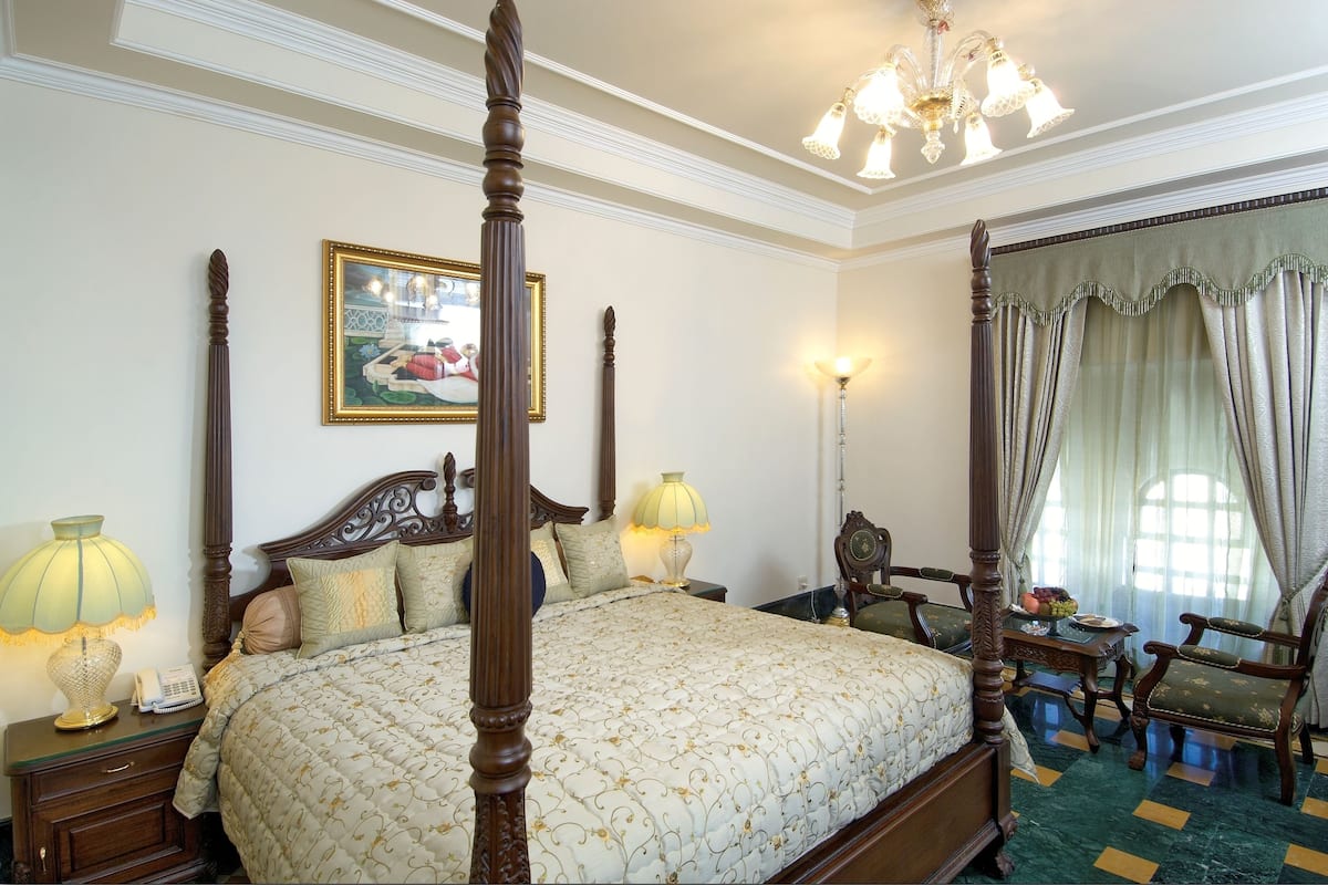 Una cama con dosel, una colcha, dos lámparas de noche, una mesita de noche, un cuadro enmarcado, un candelabro y una ventana con cortinas.