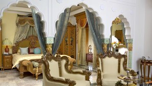 Una habitación lujosa con una cama grande, sillas ornamentadas y una mesa de comedor dispuesta con frutas y una copa de vino.