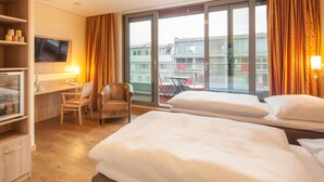 Hotelkamer met twee bedden, een bureau met stoel, een televisie en een balkon met uitzicht op gebouwen.