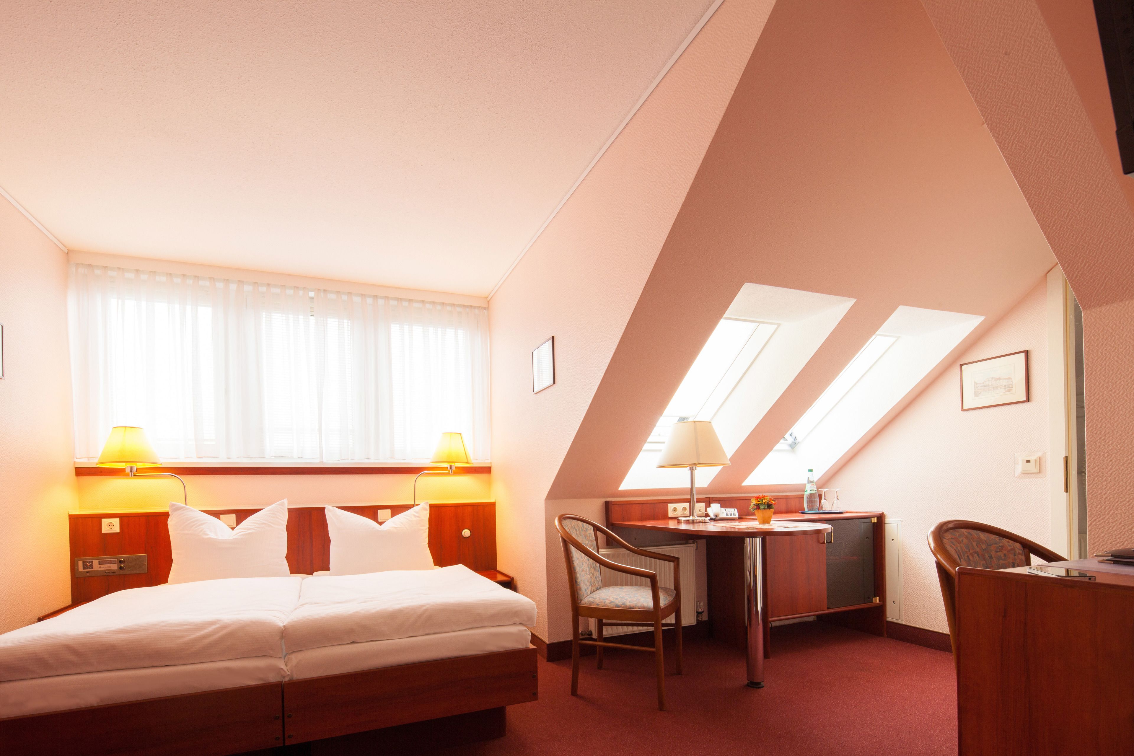 Quarto de hotel com cama, escrivaninha, cadeira e claraboia.