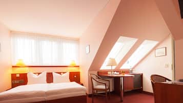 Quarto de hotel com cama, escrivaninha, cadeira e claraboia.
