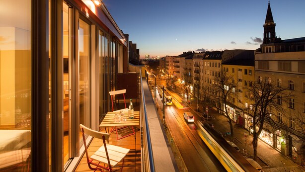 Een balkon met een tafel en stoelen, uitkijkend op een stadsstraat 's nachts, met gebouwen en een kerktoren op de achtergrond.