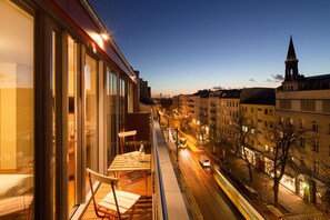 Un balcón con una mesa y sillas, con vista a una calle de la ciudad por la noche, con edificios y la aguja de una iglesia al fondo.