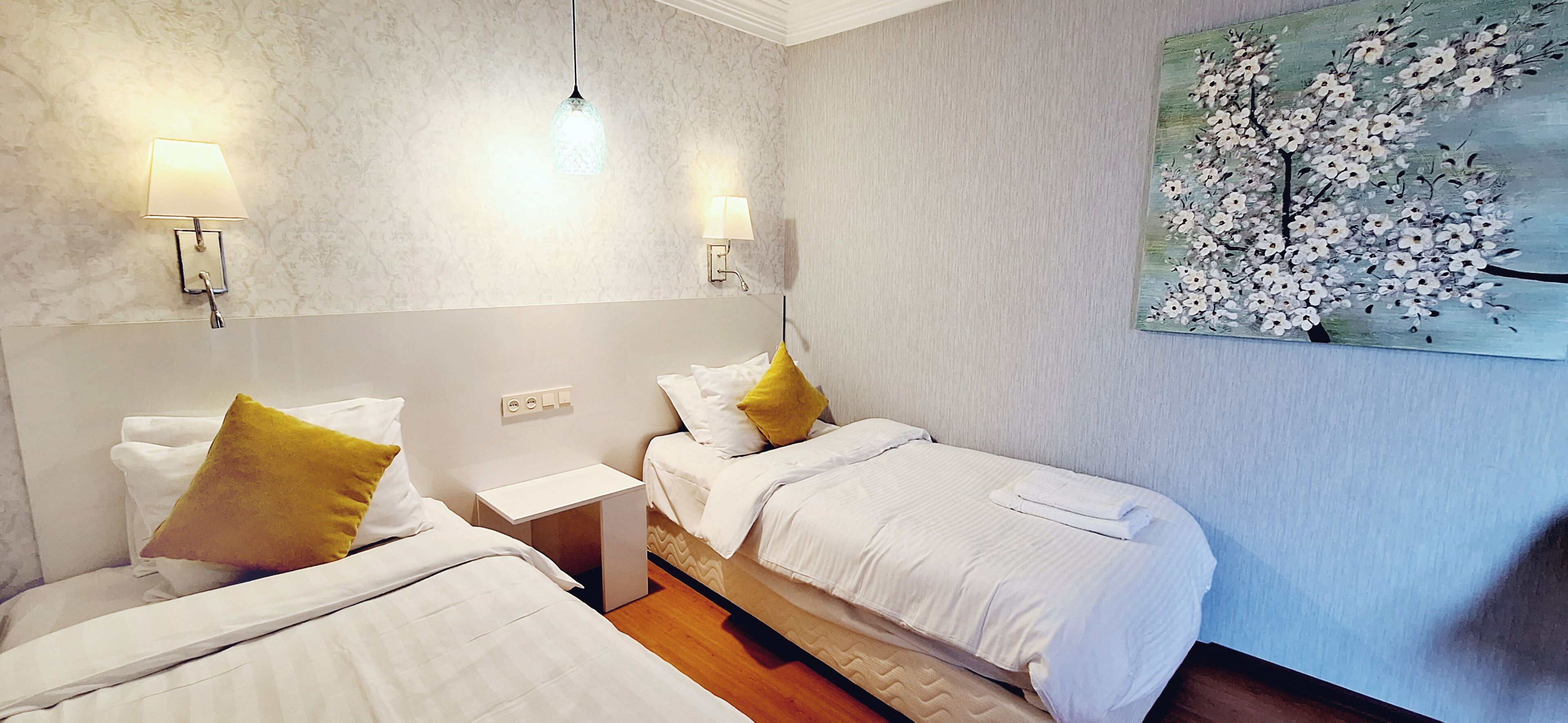 A hotel room with two beds, a nightstand, a wall-mounted lamp, and a painting of a tree.