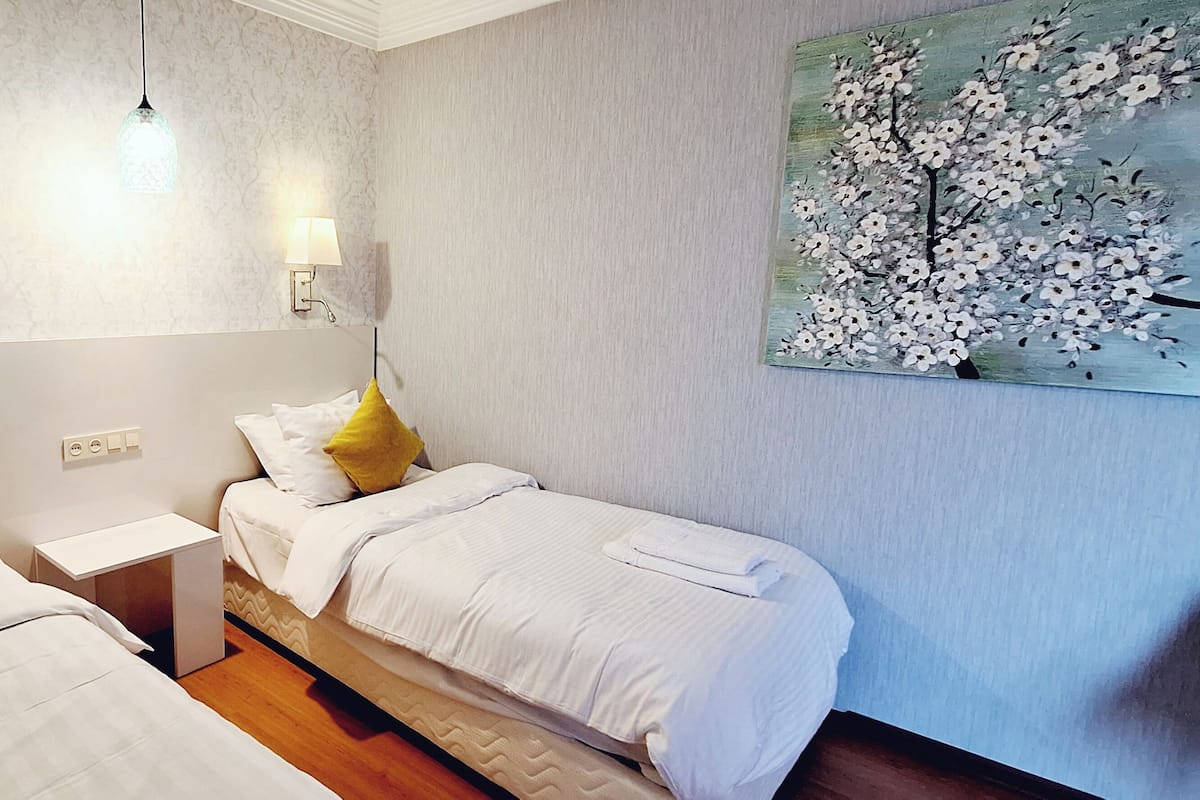 A hotel room with two beds, a nightstand, a wall-mounted lamp, and a painting of a tree.