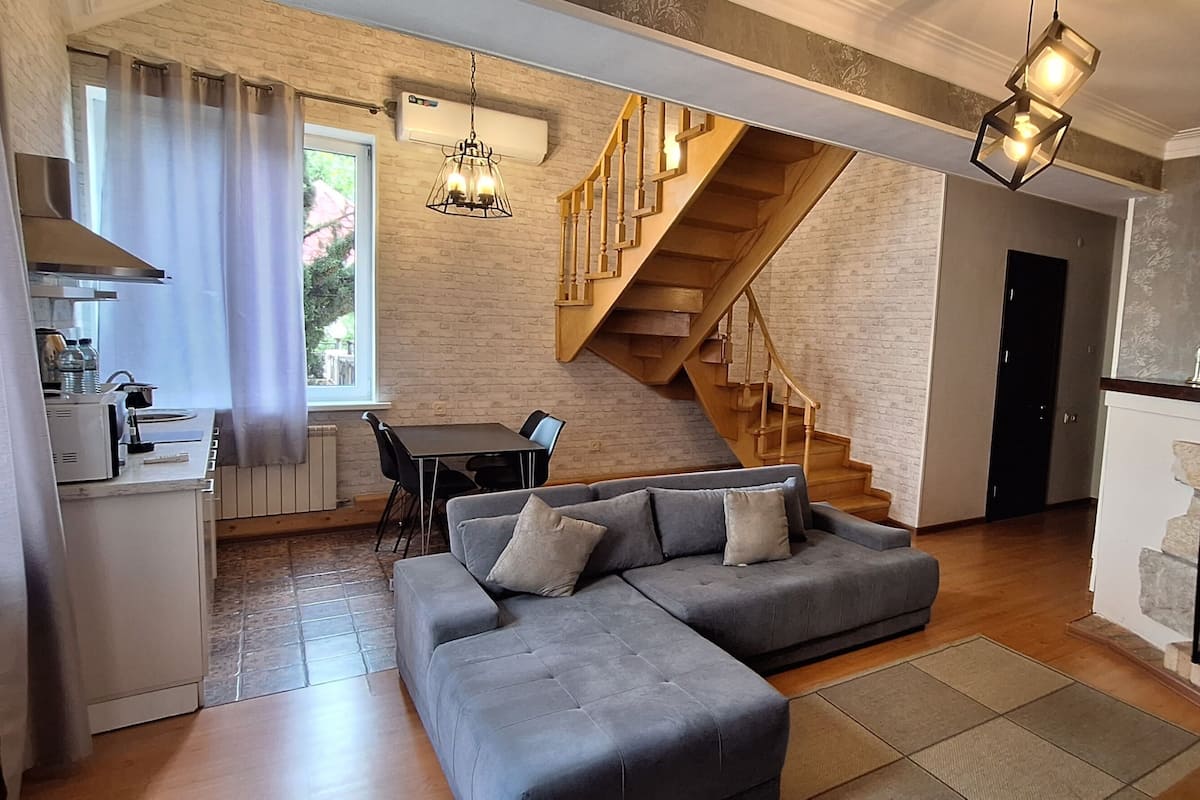A modern living room with a grey sectional sofa, a fireplace, a staircase, and a kitchen area with a dining table and chairs.