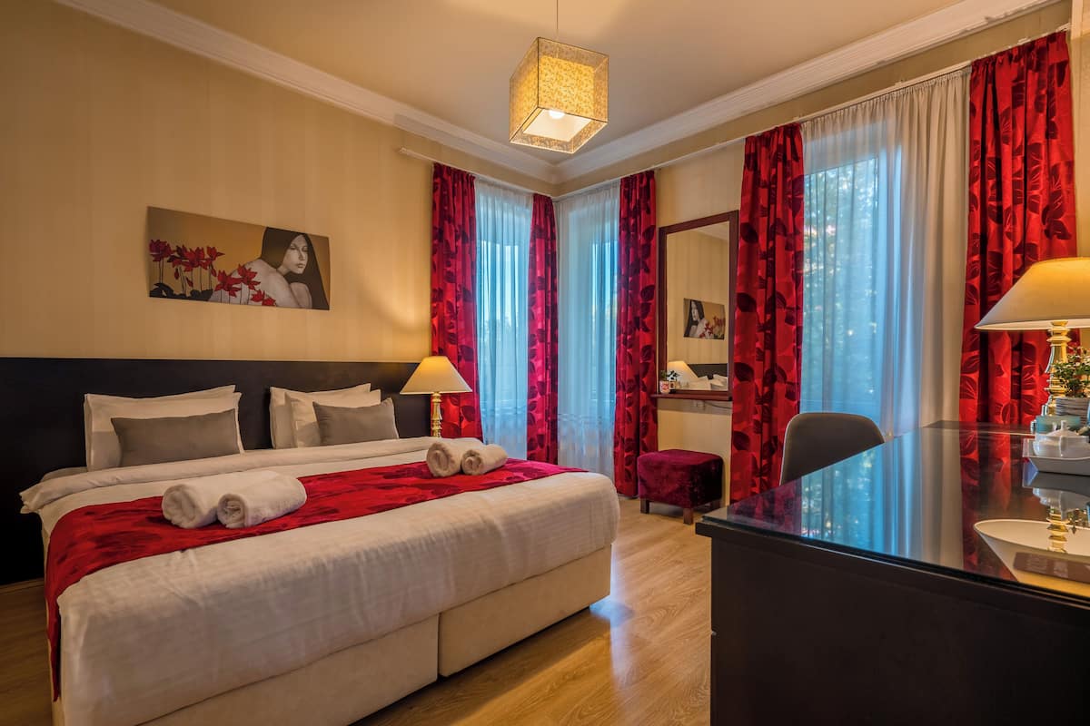 A hotel room with a large bed, two bedside lamps, a desk with a chair, and a large window with red curtains.