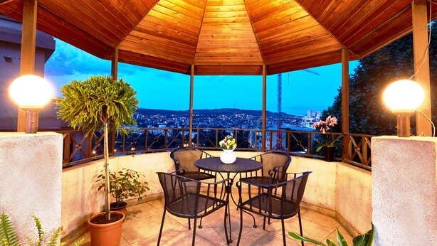 A covered patio with a wooden roof, a table with chairs, potted plants, and a view of a cityscape at dusk. - Iliani Hotel (Tbilisi)