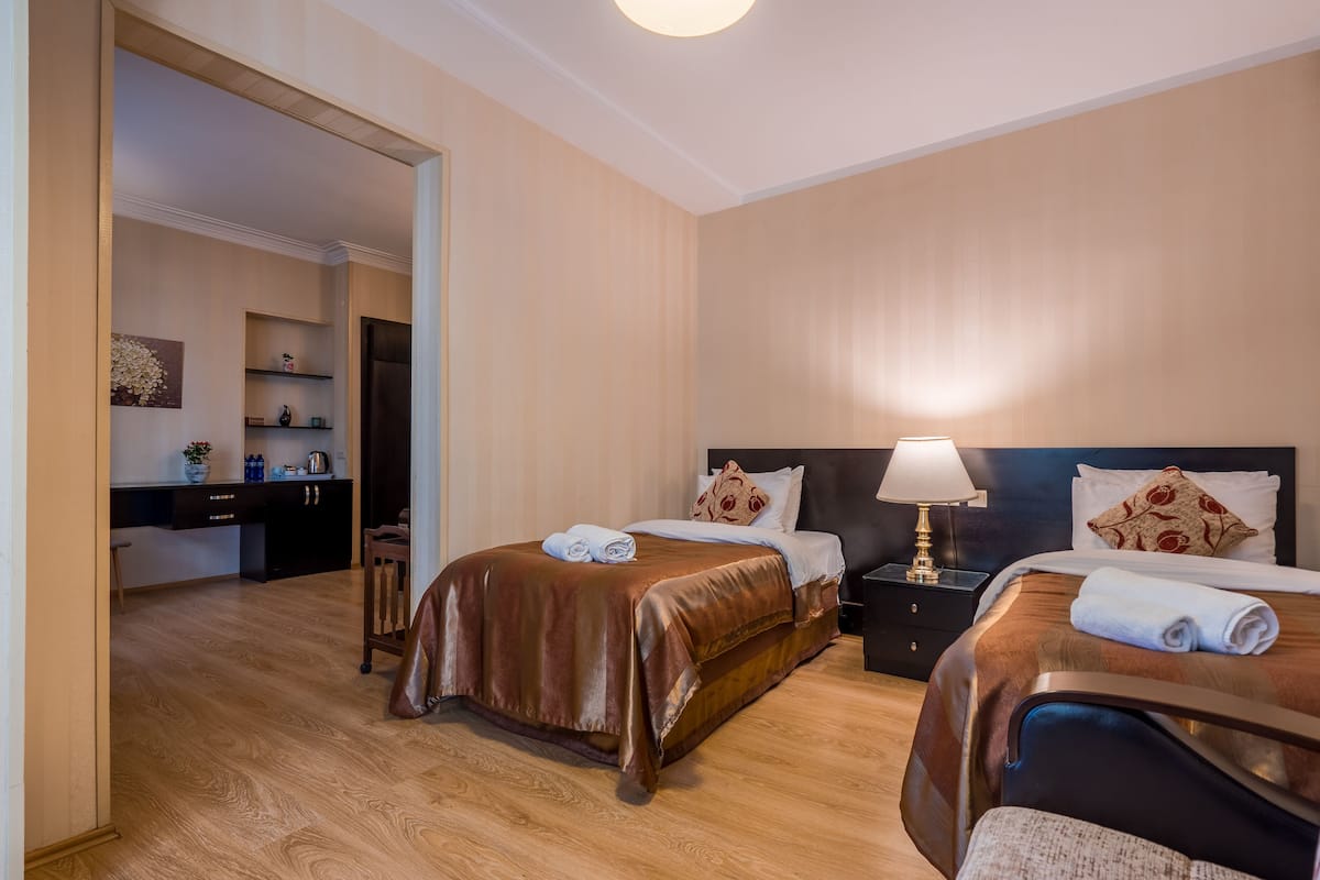 A hotel room with two beds, a nightstand with a lamp, a wooden floor, and a kitchenette in the background.