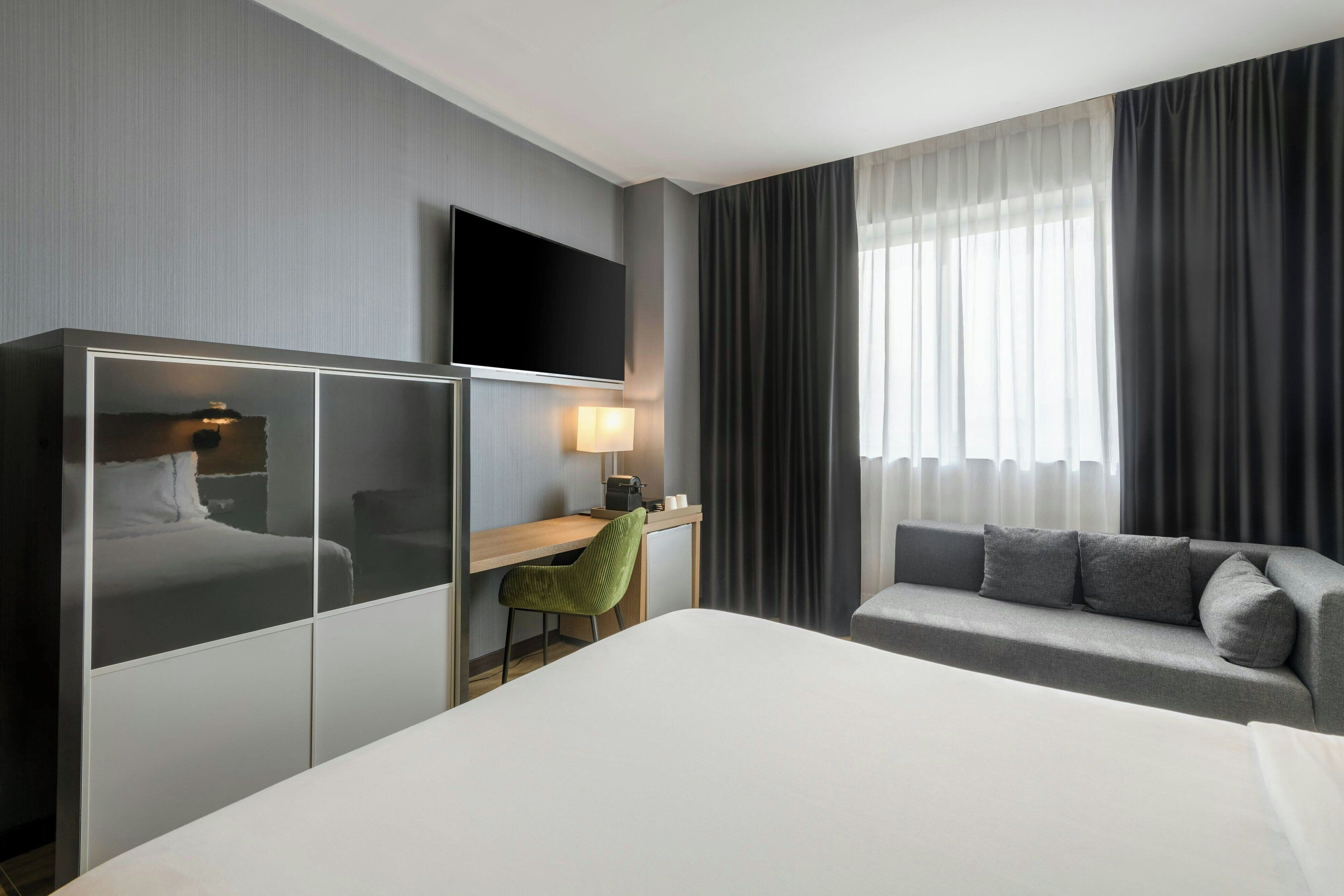 Una habitación de hotel con cama, escritorio, televisor, sofá y ventana con cortinas.