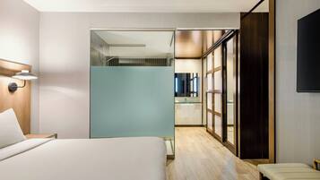 Une chambre d’hôtel moderne dotée d’un grand lit, d’un bureau avec un ordinateur portable, d’un banc, d’une cloison en verre et d’une salle de bain visible à travers une porte en verre.