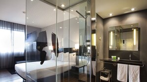 A modern hotel room with a glass-enclosed shower, a large bed, a vanity with a mirror, and a small seating area.