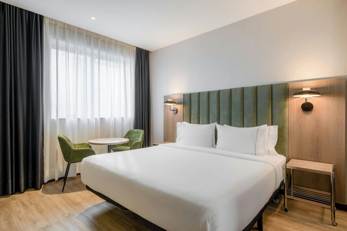 A hotel room with a large bed, a small round table, a green chair, and a window with sheer curtains.