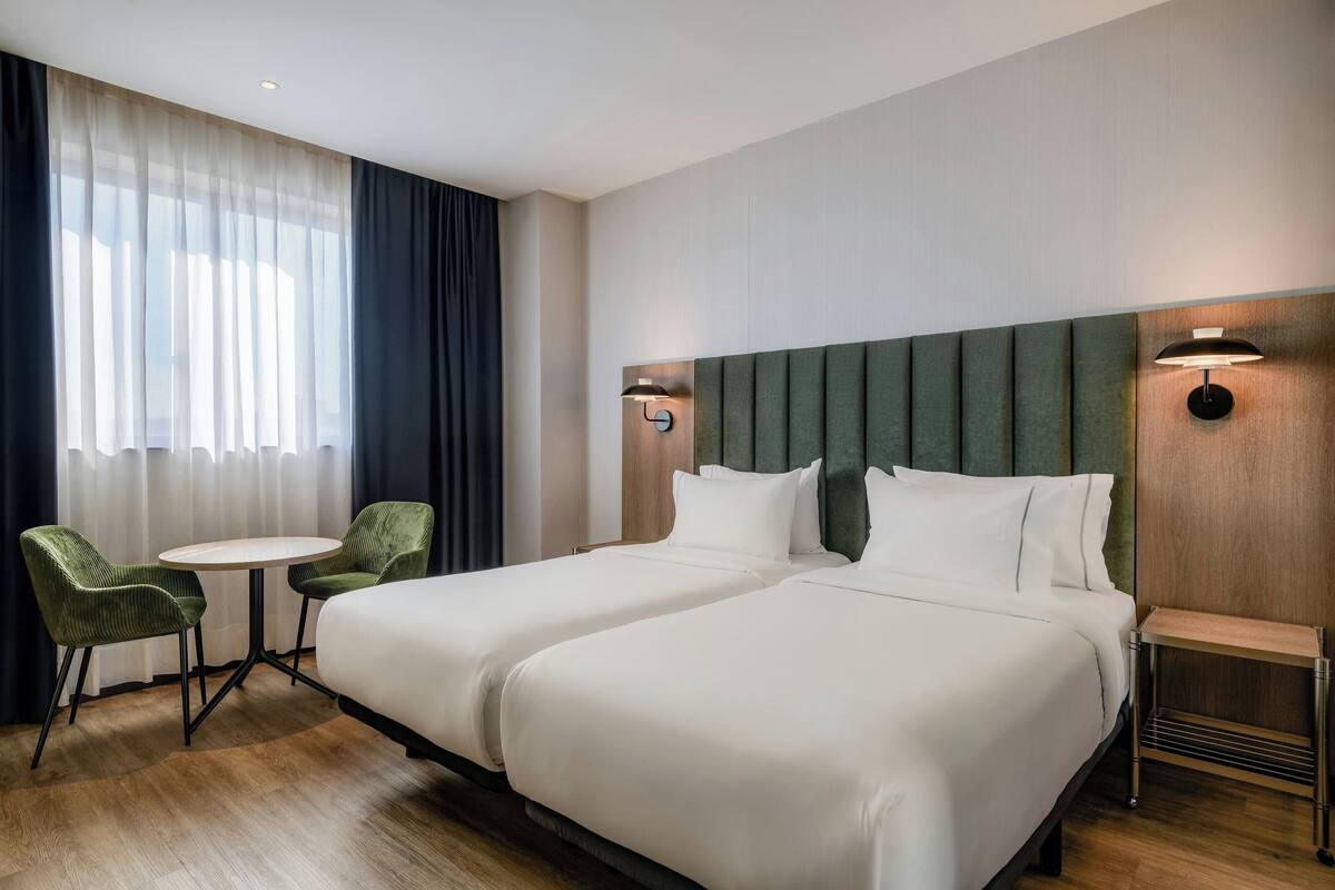 A hotel room with two beds, a small round table, two green chairs, and a window with sheer curtains.