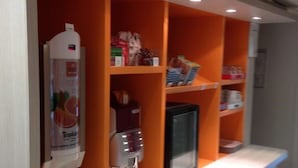A counter with a beverage dispenser, a refrigerator, and shelves with various items. - Premiere Classe Charleville Mezieres (Charleville-Mezieres)