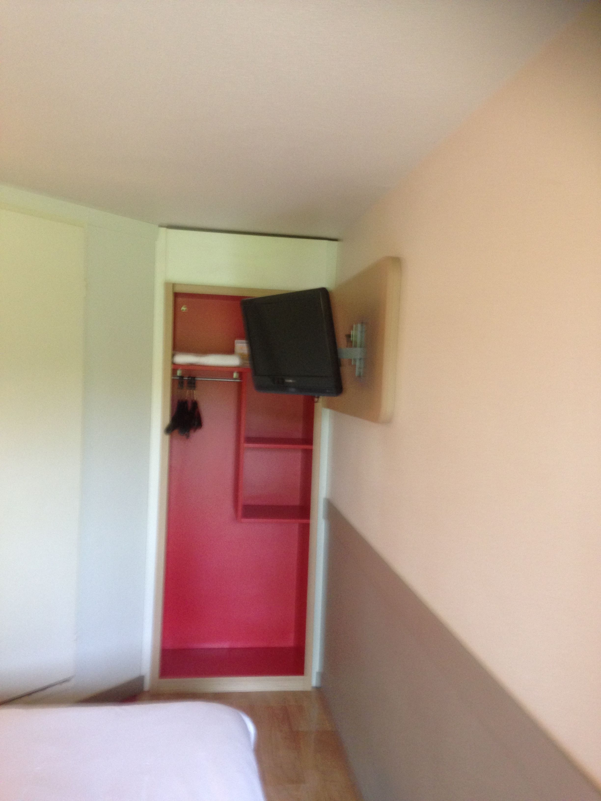 A small room with a bed, a red door, and a television mounted on the wall.